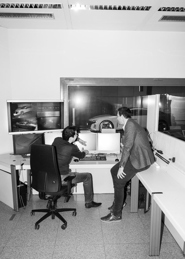 Dr. Kentaro Zens and Thomas Redenbach in discussion in front of a number of computer screens, with the Audi RS e-tron GT behind a window in the background.